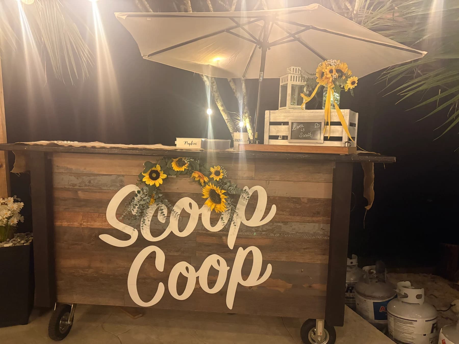 Wooden Scoop Coop event bar at twilight, dressed with sunflowers and lanterns under a market umbrella